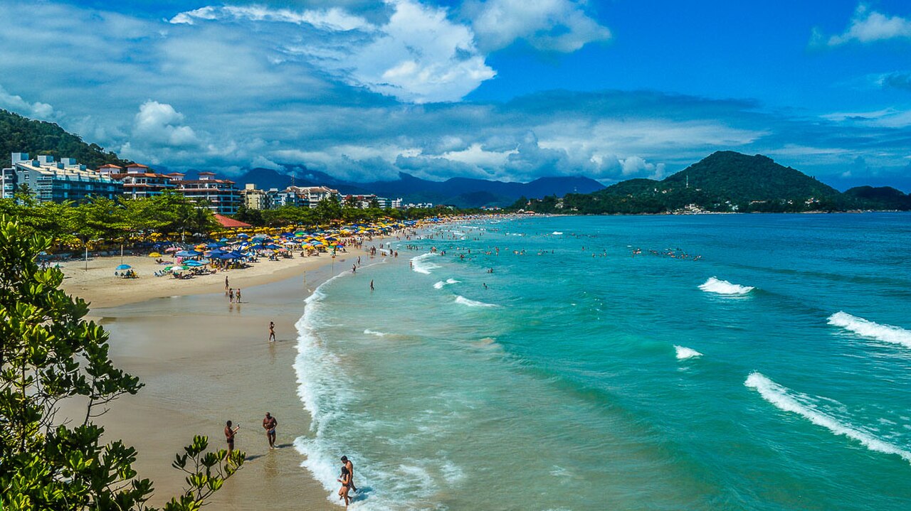Praia Grande de Ubatuba em dia de sol, com banhistas na areia e o morro ao fundo.