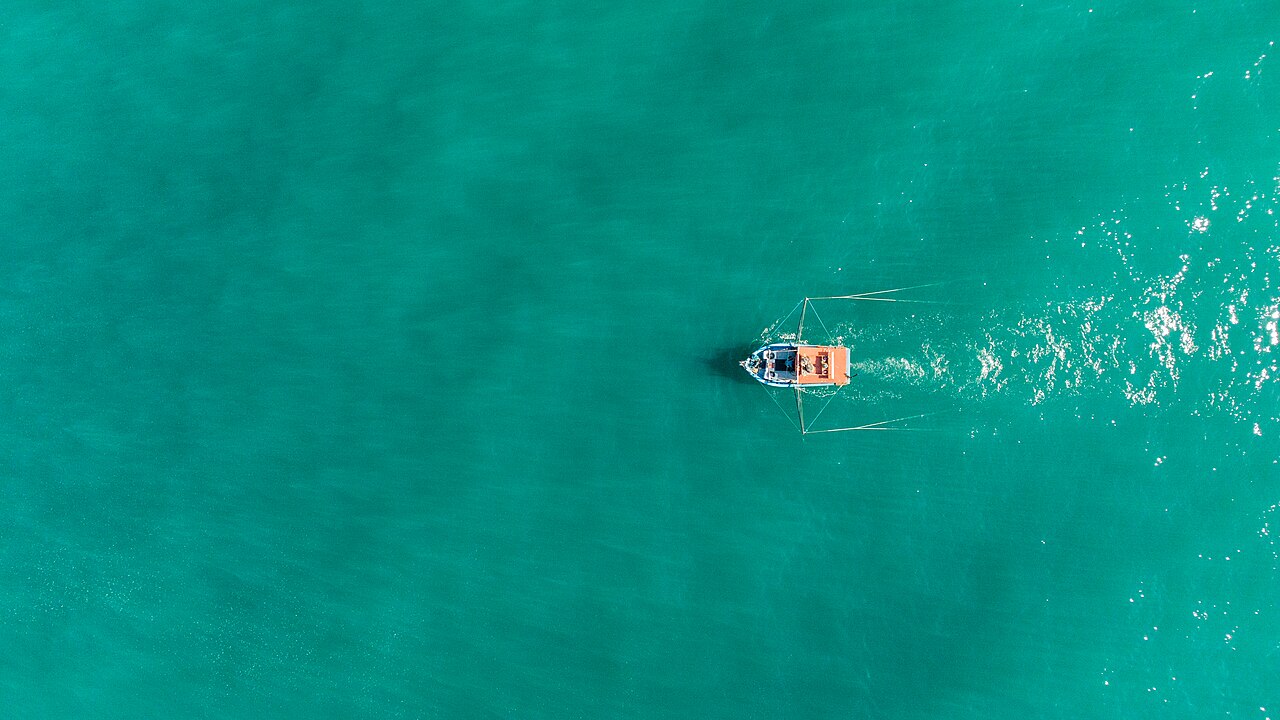 Vista aérea de um barco de pesca arrastando a rede em mar verde-esmeralda.
